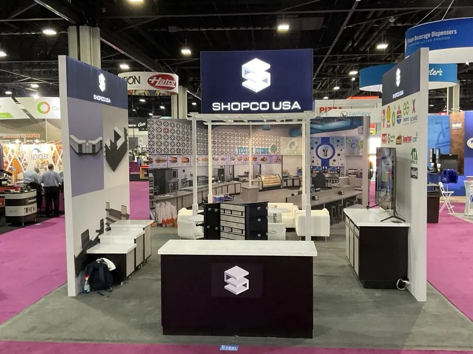 The Shopco USA exhibit on a show floor, with an overhead sign, product shelving and a branded counter.