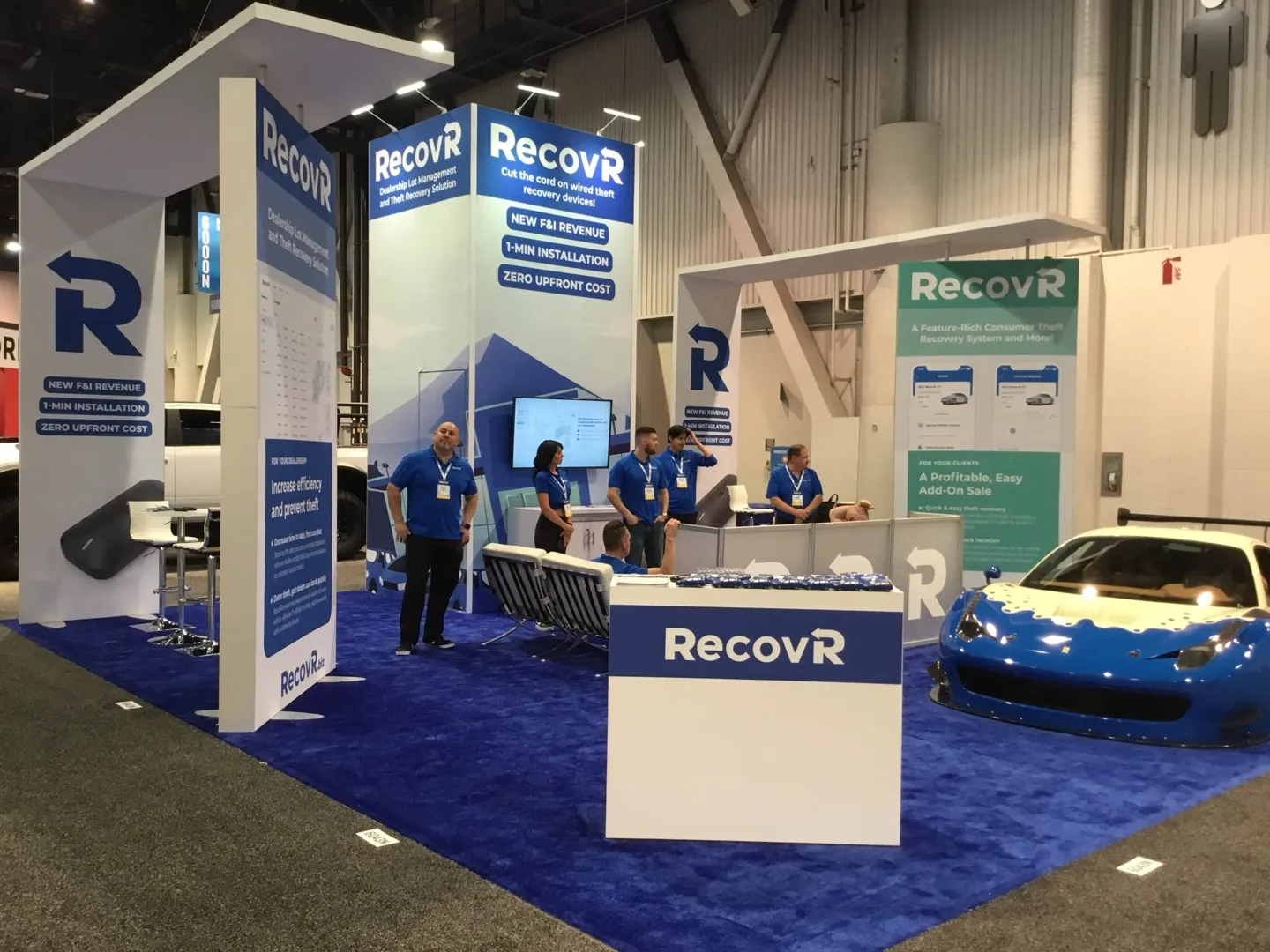The RecovR exhibit open on a show floor, staffed at the counter, with a sports car parked on the blue carpet.