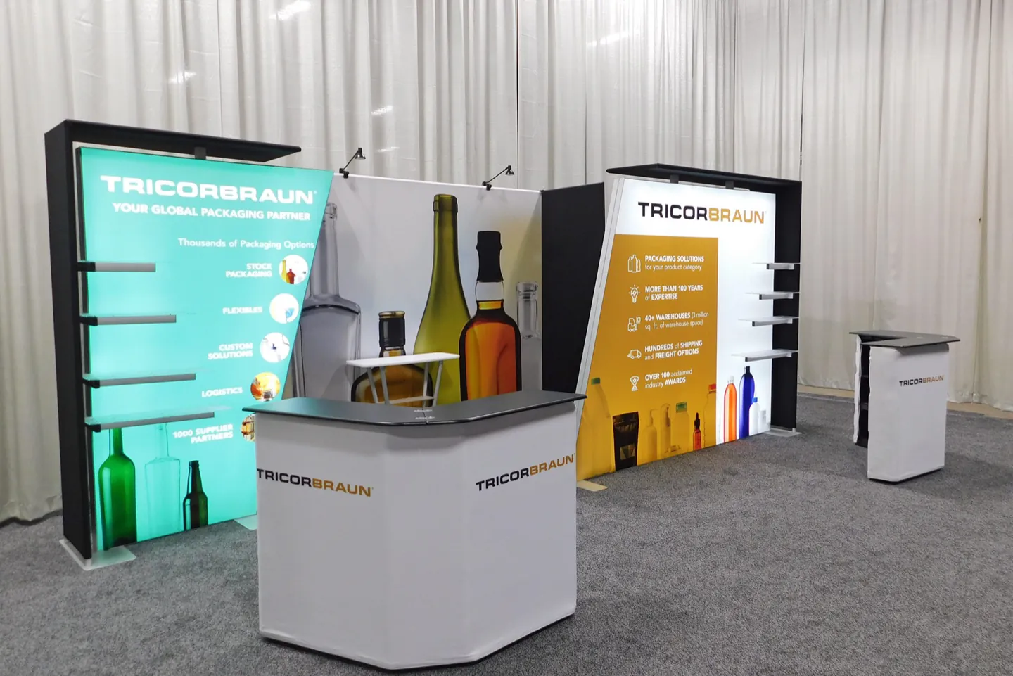 The TricorBraun exhibit on a show floor: a backlit bottle display wall and a branded counter.