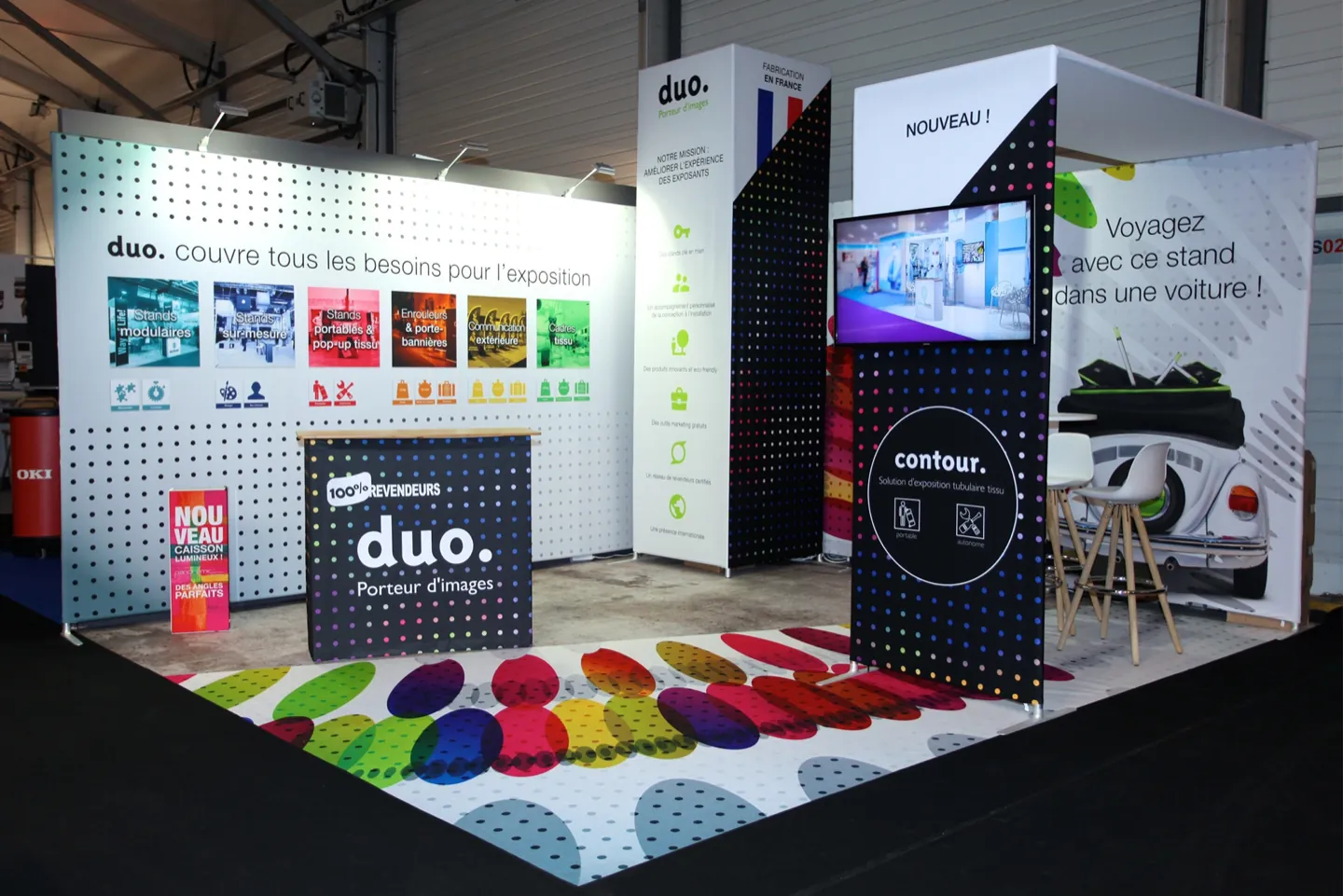 The Duo exhibit on a show floor: a printed floor graphic, a wall of product panels and a counter.